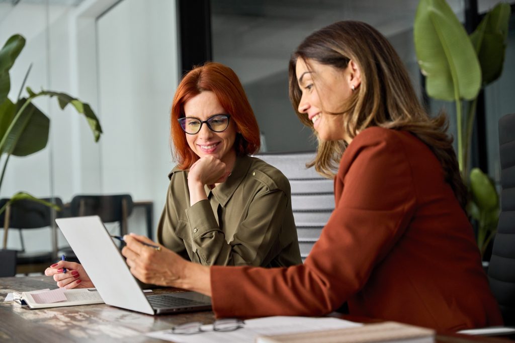 Two happy busy female coworkers working together using computer at desk in office.